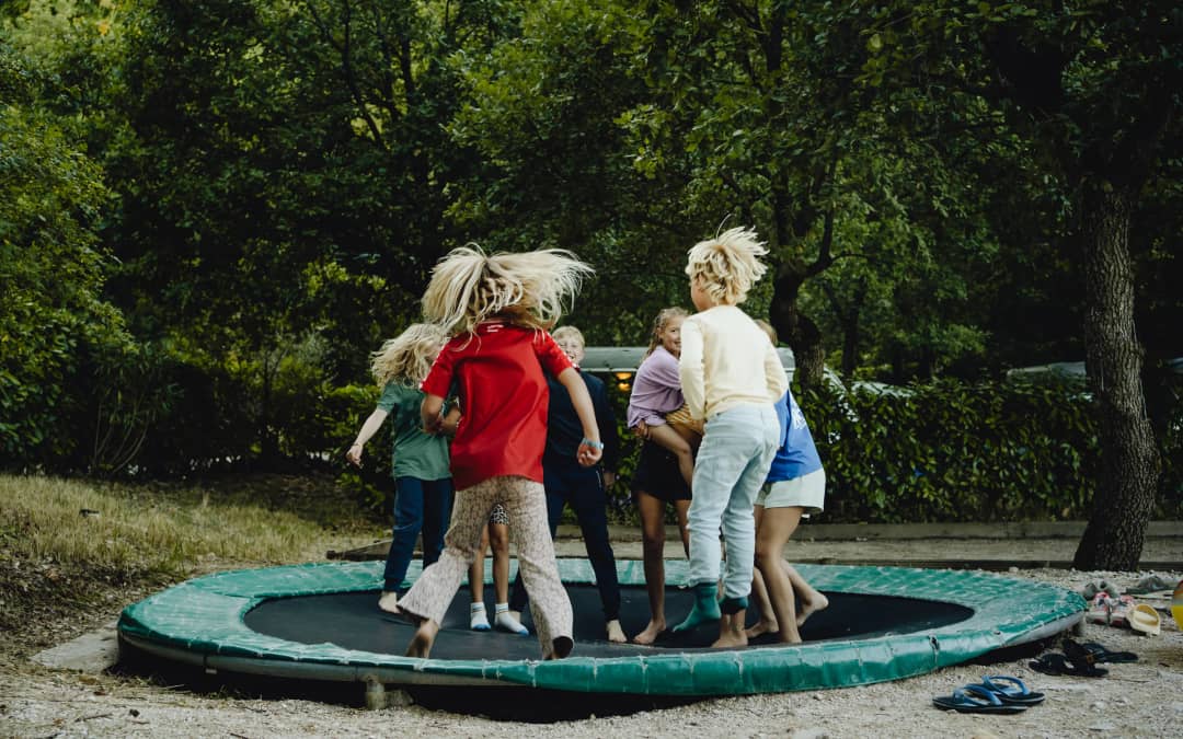 Groep kinderen springt op een ingraafde trampoline in een bosrijke camping omgeving met groen dicht op de achtergrond.