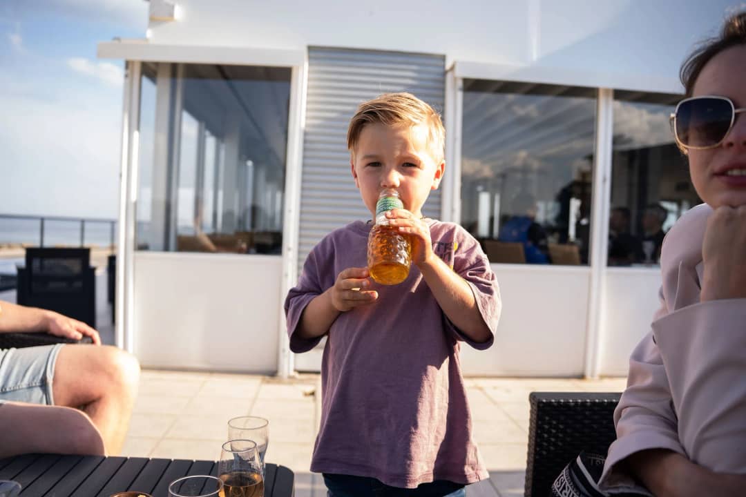 Jongen drinkt uit een flesje op een zonnig terras met glazen pui, met de zee op de achtergrond
