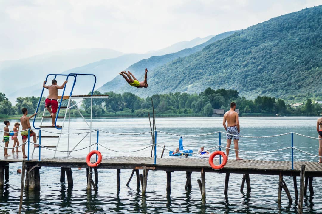 Jongen duikt van de springtoren op de steiger in het meer, met beboste bergen van Veneto op de achtergrond