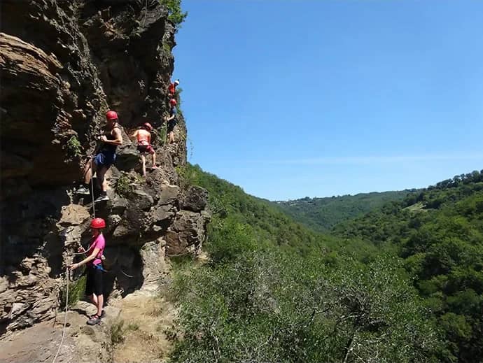 Rij klimmers met rode helmen op een via ferrata boven een groene vallei onder een strakblauwe lucht