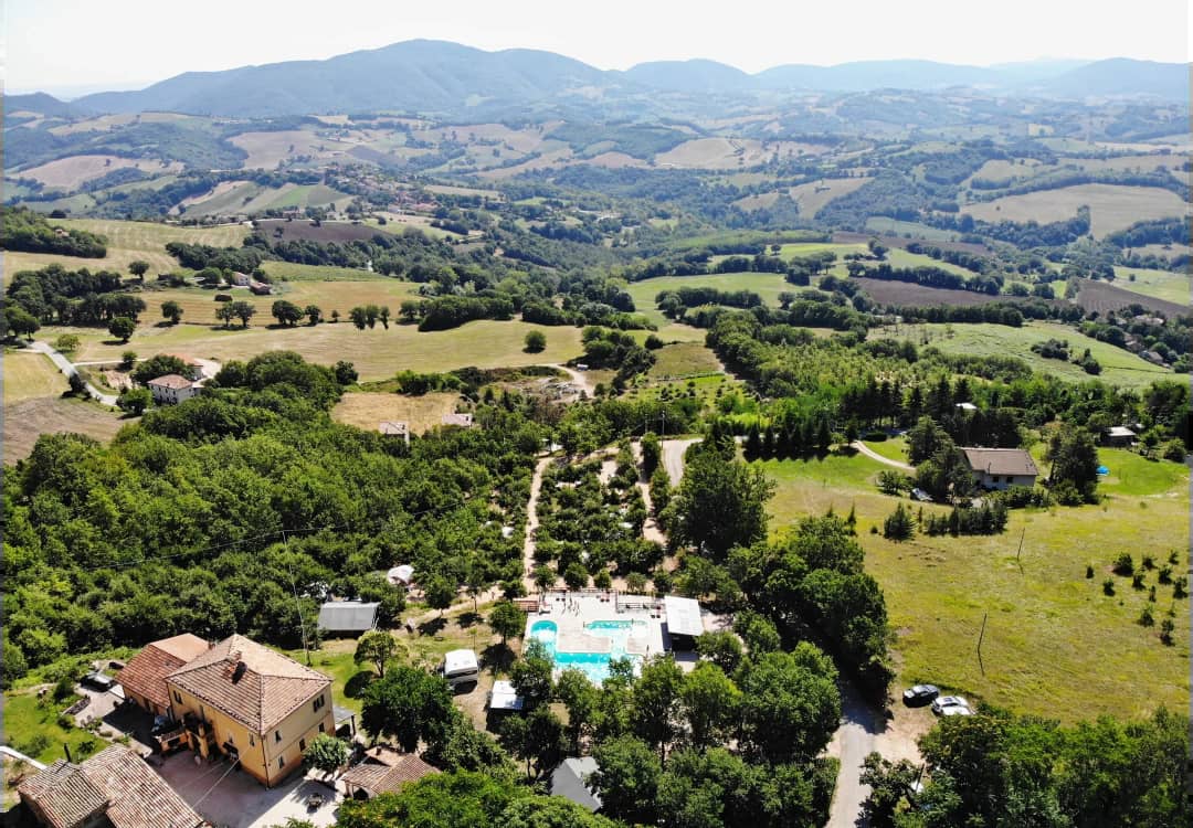 Luchtfoto van Camping Casa Tartufo in Toscaanse heuvels met zwembad, geel huis en groene velden omringd door bomen en landschap.